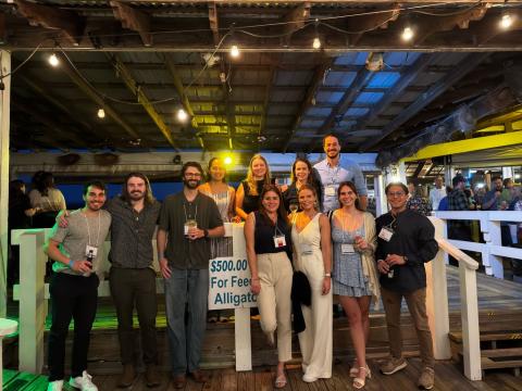 A group of students wearing lanyards and their professor smile for a group photo on a wooden dock with white wood railings and a wooden ceiling.