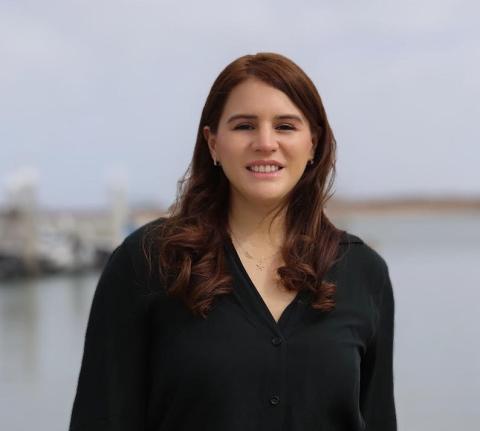 Mariana Rivera-Higueras wears a black long-sleeved blouse and stands smiling in front of an ocean shoreline.