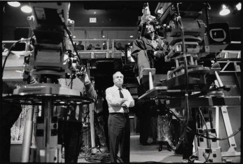 Black and white. A man in a dress shirt and tie stands with his arms folded behind two giant cameras in a television studio.