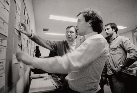Black and white. In an office, three men rearrange a cork board full of notecards.