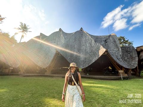 A person stands in front of a bamboo house with sloped roofs.