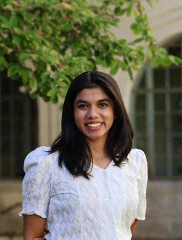 A person in a white shirt stands in front of a tree outside a building.