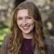 Bella Gray Bella Gray wears a burgundy shirt and a brown plaid blazer with a gold necklace. She smiles and stands in front of a blurred forest background.