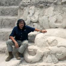 Carlos Morales-Aguilar Carlos Morales-Aguilar wears a blue button-down shirt. grey pants, and brown boots. He sits on the steps of an enormous, ancient white stone structure, with ornate curved carvings on the walls.