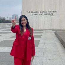 Celeste Flores Celeste Flores wears a bright red suit and makes the "hook em" sign outside of the Lyndon Baines Johnson Library and Museum, which is a windowless, gray concrete building surrounded by pavement and the Austin city skyline.