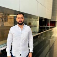 Aldo Galli Aldo Galli wears a white button-down shirt and stands on a walkway lined with glass railings. The building behind him has a floor-to-ceiling glass window, and above the window is a wall of white-paneled siding.