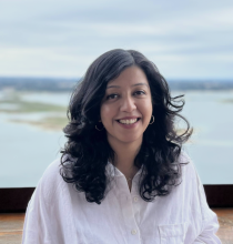 Anjali Singh Anjali Singh wears a white button-down shirt and smiles in front of a lake with some green island patches and a cloudy blue sky.