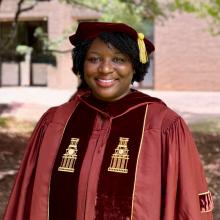 Anastazja Harris Anastazja Harris wears red convocation robes with a yellow tassel on the cap, and stands in front of a brick building.