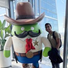 Vignesh Sella Vignesh Sella wears a black backpack and a green Oden t-shirt. He stands next to a statue of the green Android robot dressed in a cowboy costume. To the left of the robot is a plant. Behind them is a glass window overlooking Austin.