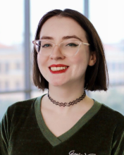 Emma Ryan Emma Ryan wears a green blouse, a black choker necklace, and clear horn-rimmed glasses. She smiles in front of a blurred background of a floor-to-ceiling window.