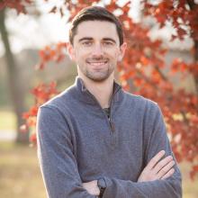 Nathan Herrmann wears a grey sweater and smiles with his arms crossed in front of a tree with orange autumn leaves.
