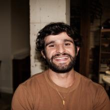 Darius wears a brown sweater and a gold necklace and smiles in front of a white pillar.