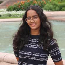 A woman with long dark hair wears glasses and a black and grey striped sweater in front of a large stone water fountain.