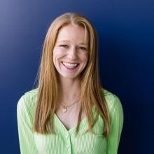 A woman wears a lime green cardigan and a gold necklace and smiles in front of a dark blue background.