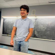 A man in a blue shirt stands in front of a blackboard.