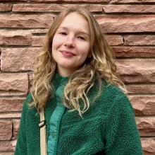 A woman smiles and wears a green sweater in front of a red stone wall.