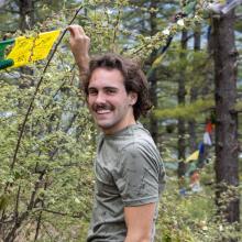 Kyle Dickens A man in a grey shirt holds up a yellow flag in front of a green bush in a forest.