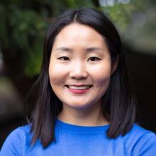 Namuun Clifford A woman in a blue blouse smiles in front of a forest background.