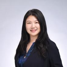 A woman wears a black blouse and smiles in front of a grey background.