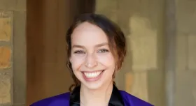 Shoshi Reich wears a purple suit and smiles in front of a brown stone wall.