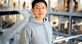 A man with black hair, grey glasses, and a grey hoodie stands in front of a metal railing. Behind him is a staircase leading to rooms lined with floor-to-ceiling windows.