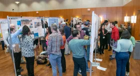 University students and professors stand in a room with wood-paneled walls and hardwood floors, all crowding posters hung on metal stands and lined in rows throughout the room. The students presenting their posters wear red lanyards.