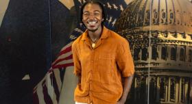 A man in an orange shirt stands in front of a mural depicting a capitol building and an American flag.