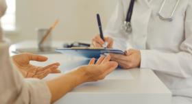A doctor in a lab coat writes notes on a tablet while speaking with a patient at a white table.