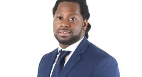 A man wears a blue suit and tie and stands in front of a white background.