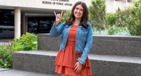 A person wears an orange dress and a jean jacket and makes a "Hook 'em Horns" hand sign in front of an academic building.