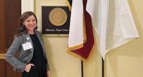 A person wears businesswear and stands next to a plaque marking the state of Texas and two flagpoles.