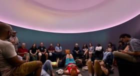 A group of students, some sitting along the wall and others reclining on the floor, observe the ceiling of a skyspace.