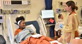 Two nursing students simulate a patient-nurse interaction in a hospital bed.