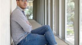 A person wears jeans and a dress shirt and sits on a padded bench in a hallway.