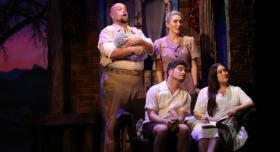 Four actors pose in 1940s era costumes onstage with the set of a brick building behind them.