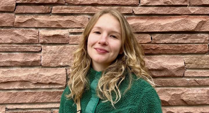 A woman wears a green sweater and smiles in front of a red stone wall.