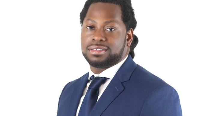 A man wears a blue suit and tie and stands in front of a white background.